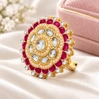 A gold-plated mandarin-style ring featuring a central clear oval stone surrounded by milky white pearls, clear round stones, and deep pinkish-red oval stones, all set in a polished gold-tone band on a white background.