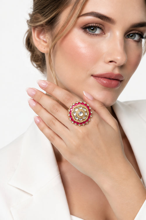 A close-up lifestyle shot of a woman wearing a large, ornate Indian-style ring with a gold-toned circular setting, a central clear stone, and rows of red and white stones. She is wearing a gold embellished traditional outfit against a warm, blurred bokeh background.