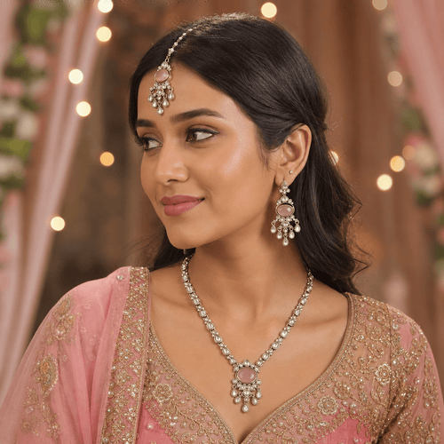 A woman wearing a traditional Indian jewelry set including a necklace, matching earrings, and a forehead jewel (maang tikka) with pink stones and silver-toned metal, paired with a pink embroidered blouse.