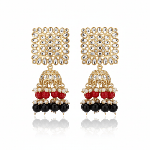 A pair of gold-plated Indian jhumka earrings with silver-toned rhinestone-embellished square studs and dangling bell-shaped charms featuring red, white, and black beads, displayed on a dark wood background.