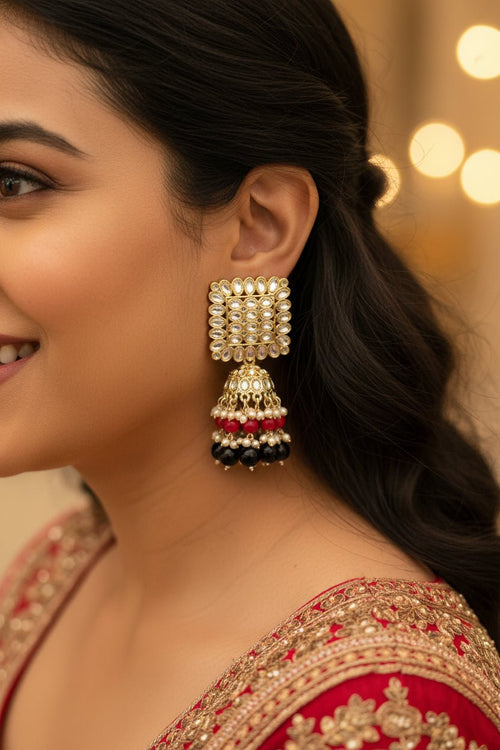 A close-up of a woman wearing a large gold-plated traditional Indian jhumka earring adorned with white stones, red, and black beads, paired with a red embroidered blouse.