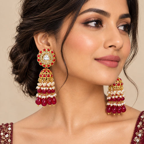 A pair of gold-plated Indian jhumka earrings decorated with red stones, white faux pearls, and a central clear triangular crystal on a white background.