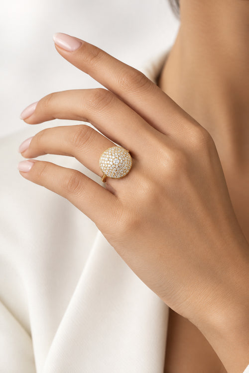 A close-up of a hand wearing a large, round, textured gold ring with a floral or sunburst pattern, set against a neutral, light-textured background.