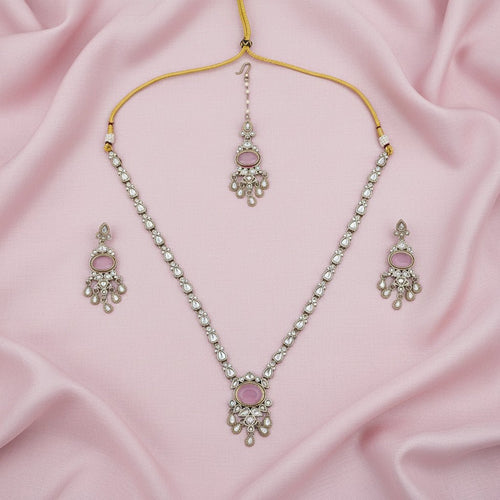 A silver-toned jewelry set featuring a necklace, matching earrings, and a dangling maang tikka, all adorned with pink stones and small clear stones, displayed on a white background with loose matching stone embellishments arranged around them.