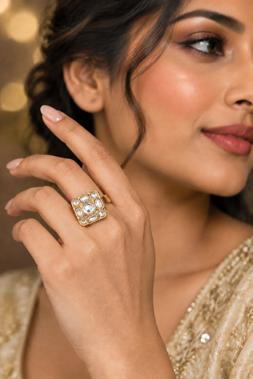A gold-toned square ring with clear stones, worn on a person's hand against a light textured wall.