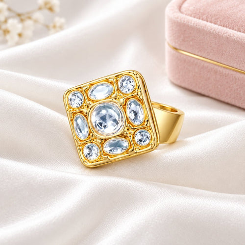 A gold square ring featuring a central round light blue gemstone surrounded by eight smaller light blue oval and round stones, set in a polished yellow gold bezel on a white background.