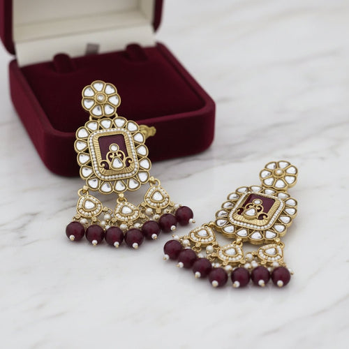 A pair of ornate gold-plated Indian jhumka earrings decorated with white faux pearls, a central maroon rectangular inlay, and dangling maroon beads at the bottom, shown on a white background with a soft reflection.