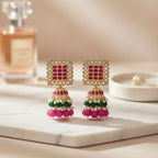 A pair of gold-plated Indian jhumka earrings with pink and green stones, displayed on clear plastic stands against a textured white background.