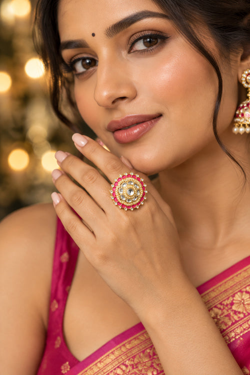 A close-up lifestyle shot of a woman wearing a large, ornate Indian-style ring with a gold-toned circular setting, a central clear stone, and rows of red and white stones. She is wearing a gold embellished traditional outfit against a warm, blurred bokeh background.